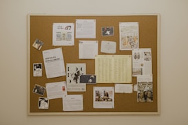 A cork bulletin board attached to a wall, filled with various documents, images, and flyers pinned. The contents include photos, printed text pages, graphic illustrations, and lists. Several sheets are loosely organized and thumbtacked to the board.