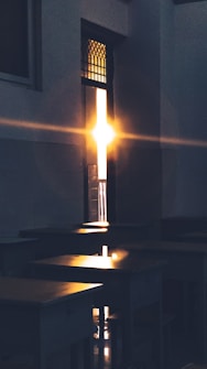 Sunlight streams through a partially open door, casting warm, glowing light across the desks of an empty classroom. The room is dimly lit, emphasizing the contrast with the bright sunlight, which creates a peaceful and serene atmosphere. The architecture features a tall doorway with intricate glass panes at the top.