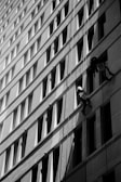 Two workers are suspended on ropes, cleaning the windows of a tall building. The geometric pattern of the windows creates a dynamic visual effect in black and white. The sunlight casts shadows that add depth to the image.
