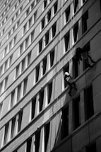 Two workers are suspended on ropes, cleaning the windows of a tall building. The geometric pattern of the windows creates a dynamic visual effect in black and white. The sunlight casts shadows that add depth to the image.