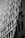 Two workers are suspended on ropes, cleaning the windows of a tall building. The geometric pattern of the windows creates a dynamic visual effect in black and white. The sunlight casts shadows that add depth to the image.