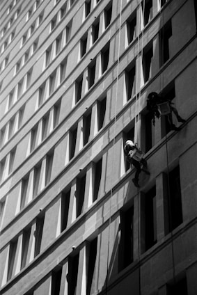 Two workers are suspended on ropes, cleaning the windows of a tall building. The geometric pattern of the windows creates a dynamic visual effect in black and white. The sunlight casts shadows that add depth to the image.