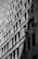 Two workers are suspended on ropes, cleaning the windows of a tall building. The geometric pattern of the windows creates a dynamic visual effect in black and white. The sunlight casts shadows that add depth to the image.