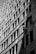 Two workers are suspended on ropes, cleaning the windows of a tall building. The geometric pattern of the windows creates a dynamic visual effect in black and white. The sunlight casts shadows that add depth to the image.