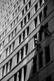 Two workers are suspended on ropes, cleaning the windows of a tall building. The geometric pattern of the windows creates a dynamic visual effect in black and white. The sunlight casts shadows that add depth to the image.