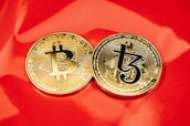 Two gold coins are placed on a glossy red surface, featuring the symbols for Bitcoin and Tezos. The coins are intricately designed with digital circuit patterns, symbolizing their status as cryptocurrencies.