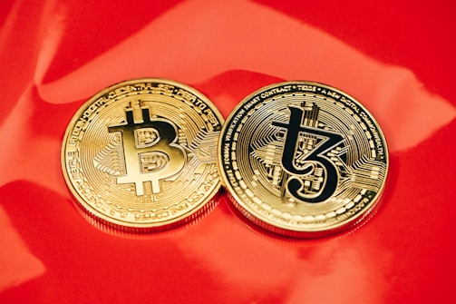 Two gold coins are placed on a glossy red surface, featuring the symbols for Bitcoin and Tezos. The coins are intricately designed with digital circuit patterns, symbolizing their status as cryptocurrencies.