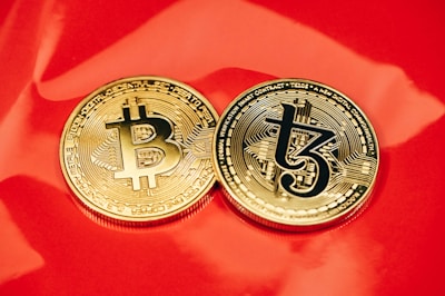 Two gold coins are placed on a glossy red surface, featuring the symbols for Bitcoin and Tezos. The coins are intricately designed with digital circuit patterns, symbolizing their status as cryptocurrencies.