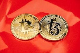 Two gold coins are placed on a glossy red surface, featuring the symbols for Bitcoin and Tezos. The coins are intricately designed with digital circuit patterns, symbolizing their status as cryptocurrencies.