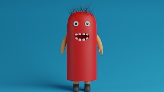 A whimsical, cartoonish character with a cylindrical, red body resembling a hot dog stands against a blue background. It has googly eyes, a wide open mouth with a few white teeth, spiky black hair, and tan-colored arms and brown shoes.