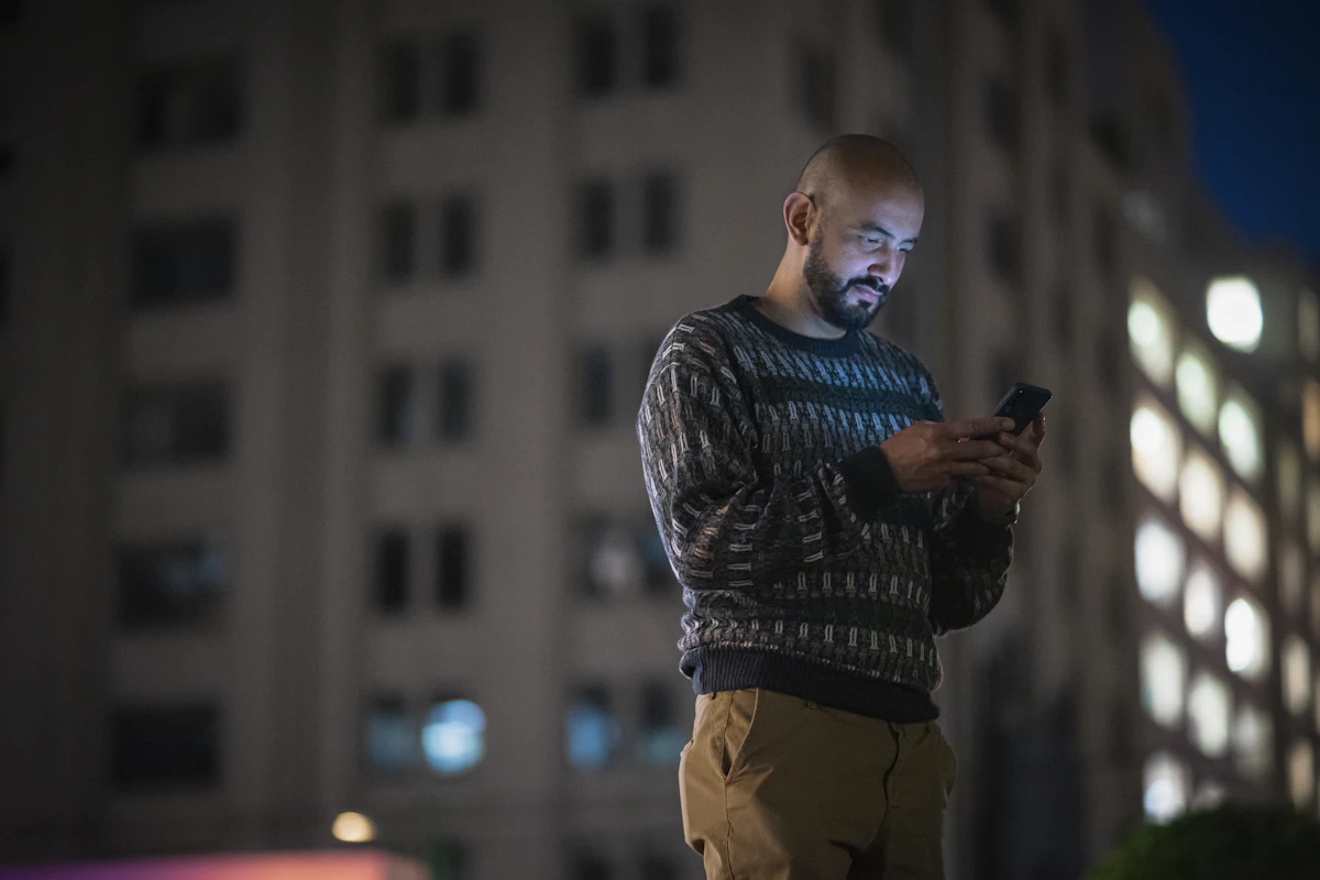 Un homme consulte son smartphone le soir, debout dans la rue devant la façade éclairée d'un immeuble