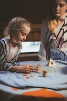 A young girl and a woman engage in a creative activity, playing with small clay sculptures on a tablecloth decorated with colorful dots. The girl is focused on the figures, which include two animal-shaped sculptures and a triangular object with a handprint design.