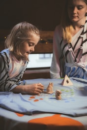 A young girl and a woman engage in a creative activity, playing with small clay sculptures on a tablecloth decorated with colorful dots. The girl is focused on the figures, which include two animal-shaped sculptures and a triangular object with a handprint design.