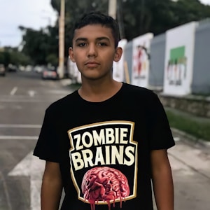 A person stands outdoors wearing a black T-shirt with a graphic design featuring the text 'Zombie Brains' above a stylized image of a brain. The background includes a blurred view of a street with trees and some painted walls.