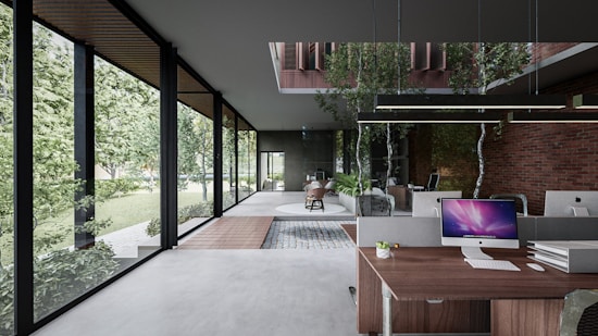 A modern office space with large glass windows providing a view of lush greenery outside. The interior features a mix of contemporary and natural elements, including brick walls and indoor plants. Desks are equipped with computer monitors and stationery. A wooden floor section adds warmth to the sleek, minimalistic design. Abundant natural light enhances the open and airy atmosphere.
