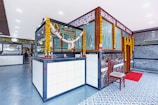 A modern and clean interior space with a cash counter adorned with floral garlands. The area features a tiled floor with a combination of blue and white patterns. A wooden chair is placed beside the counter, and there is a red mat in front of a door. Decorative panels with intricate designs are on the walls, and various signages are visible.