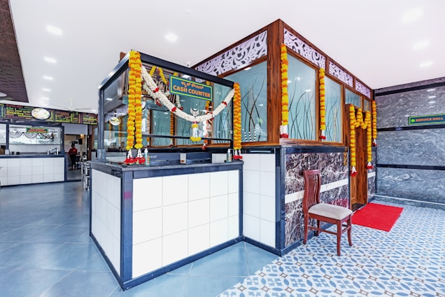 A modern and clean interior space with a cash counter adorned with floral garlands. The area features a tiled floor with a combination of blue and white patterns. A wooden chair is placed beside the counter, and there is a red mat in front of a door. Decorative panels with intricate designs are on the walls, and various signages are visible.