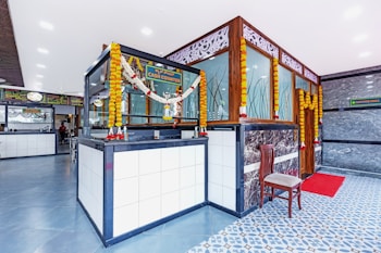 A modern and clean interior space with a cash counter adorned with floral garlands. The area features a tiled floor with a combination of blue and white patterns. A wooden chair is placed beside the counter, and there is a red mat in front of a door. Decorative panels with intricate designs are on the walls, and various signages are visible.