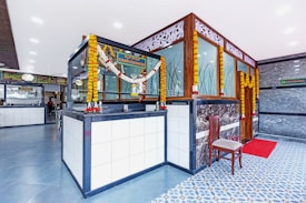 A modern and clean interior space with a cash counter adorned with floral garlands. The area features a tiled floor with a combination of blue and white patterns. A wooden chair is placed beside the counter, and there is a red mat in front of a door. Decorative panels with intricate designs are on the walls, and various signages are visible.
