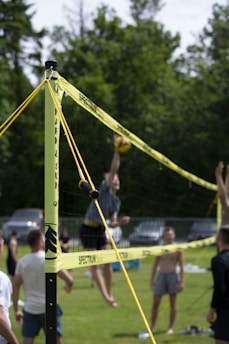 A group of people engaged in a game of volleyball on a grassy field. The net is prominently shown with yellow bands securing it. Several players are in mid-action, with one jumping to hit the ball, indicating a lively and active scene. Background features include trees and parked cars, suggesting an outdoor recreational setting.