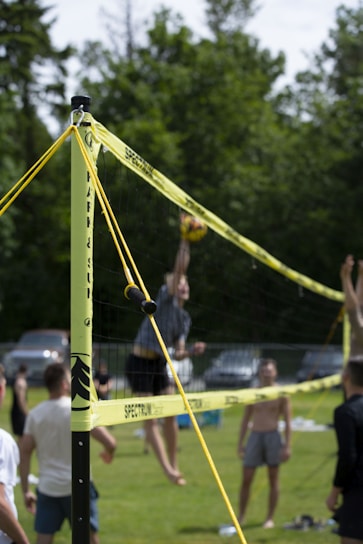 A group of people engaged in a game of volleyball on a grassy field. The net is prominently shown with yellow bands securing it. Several players are in mid-action, with one jumping to hit the ball, indicating a lively and active scene. Background features include trees and parked cars, suggesting an outdoor recreational setting.