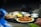 A tray is being held by a person wearing a green sleeve, showcasing an assortment of Indian dishes. The tray includes a metal bowl with a richly garnished curry, a plate of dry-fried food with herbs, and several small metal bowls containing various accompaniments. The background is slightly blurred, indicating a focus on the food.