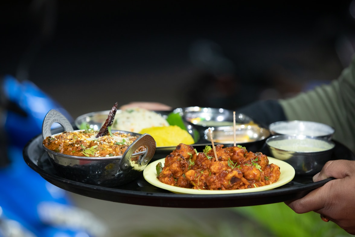 A tray is being held by a person wearing a green sleeve, showcasing an assortment of Indian dishes. The tray includes a metal bowl with a richly garnished curry, a plate of dry-fried food with herbs, and several small metal bowls containing various accompaniments. The background is slightly blurred, indicating a focus on the food.