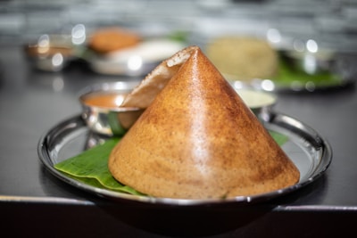 A crispy, cone-shaped dosa is placed on a stainless steel plate lined with a banana leaf. Accompanying the dosa are small bowls containing different chutneys.