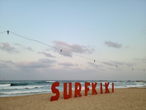 Large red letters spelling out 'SURFKIKI' are positioned on a sandy beach with gentle waves visible in the ocean. The sky is partly cloudy with a soft, muted appearance. Overhead, a string of light bulbs hangs, adding a decorative element to the scene.