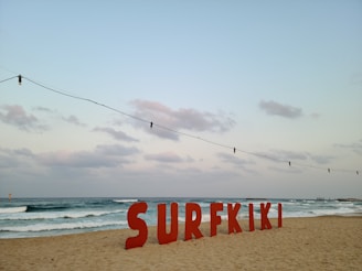 Large red letters spelling out 'SURFKIKI' are positioned on a sandy beach with gentle waves visible in the ocean. The sky is partly cloudy with a soft, muted appearance. Overhead, a string of light bulbs hangs, adding a decorative element to the scene.