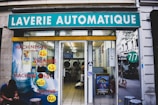 A laundromat with the sign 'Laverie Automatique' above the entrance. The glass windows display advertisements for washing and drying machines with prices. Inside, washing machines are visible along with tiled flooring. A person is seated on the left side outside, using a phone.