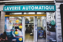 A laundromat with the sign 'Laverie Automatique' above the entrance. The glass windows display advertisements for washing and drying machines with prices. Inside, washing machines are visible along with tiled flooring. A person is seated on the left side outside, using a phone.