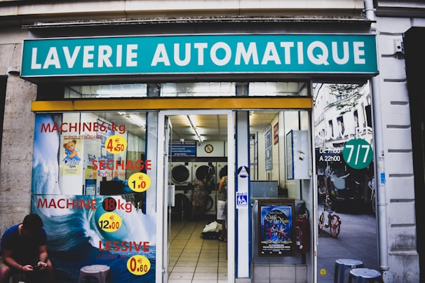 A laundromat with the sign 'Laverie Automatique' above the entrance. The glass windows display advertisements for washing and drying machines with prices. Inside, washing machines are visible along with tiled flooring. A person is seated on the left side outside, using a phone.