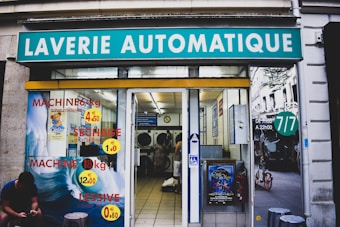 A laundromat with the sign 'Laverie Automatique' above the entrance. The glass windows display advertisements for washing and drying machines with prices. Inside, washing machines are visible along with tiled flooring. A person is seated on the left side outside, using a phone.