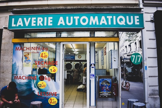 A laundromat with the sign 'Laverie Automatique' above the entrance. The glass windows display advertisements for washing and drying machines with prices. Inside, washing machines are visible along with tiled flooring. A person is seated on the left side outside, using a phone.