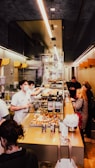 Inside a bakery, staff members wearing white uniforms and masks work behind a counter filled with pastries and wine bottles. Customers stand at the counter, examining the items and placing orders. The space is narrow and brightly lit, with a modern design featuring glass and muted colors.