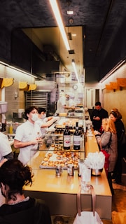 Inside a bakery, staff members wearing white uniforms and masks work behind a counter filled with pastries and wine bottles. Customers stand at the counter, examining the items and placing orders. The space is narrow and brightly lit, with a modern design featuring glass and muted colors.