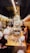 Inside a bakery, staff members wearing white uniforms and masks work behind a counter filled with pastries and wine bottles. Customers stand at the counter, examining the items and placing orders. The space is narrow and brightly lit, with a modern design featuring glass and muted colors.