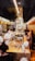 Inside a bakery, staff members wearing white uniforms and masks work behind a counter filled with pastries and wine bottles. Customers stand at the counter, examining the items and placing orders. The space is narrow and brightly lit, with a modern design featuring glass and muted colors.
