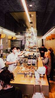 Inside a bakery, staff members wearing white uniforms and masks work behind a counter filled with pastries and wine bottles. Customers stand at the counter, examining the items and placing orders. The space is narrow and brightly lit, with a modern design featuring glass and muted colors.