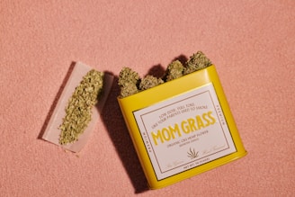 A yellow tin labeled 'Mom Grass' containing organic CBG hemp flower is open, displaying its contents. Beside it, a rolling paper partially filled with ground hemp is visible. The background is a textured, light pink surface.