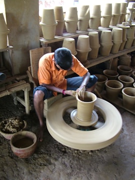 A person wearing an orange shirt and shorts is sitting on a stool, shaping a clay pot on a spinning pottery wheel. The workshop is filled with shelves stacked with finished clay pots of various sizes. The floor is covered in clay residue, and there are several containers with more clay material nearby.