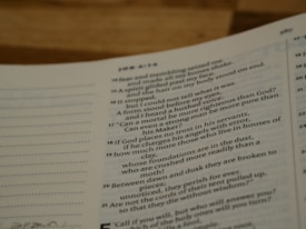 A close-up shot of an open book, likely a Bible or similar text, with several verses visible. The text is in two columns and features chapter and verse numbers. The focus is on a particular passage with the numbers appearing at the side of each line.