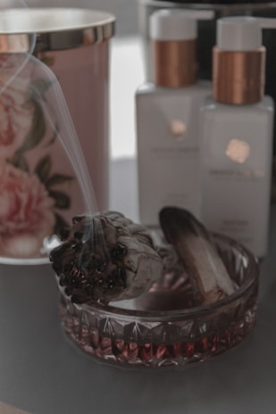 A decorative setting features a smoldering sage bundle resting in an ornate glass dish, alongside elegant bottles with white labels and rose gold accents. In the background, there is a floral-patterned container.