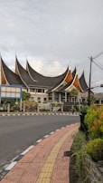 An architectural building with distinctively pointed, multi-tiered roofs resembling traditional Minangkabau architecture. It is situated at a corner with a curved road and a pedestrian pathway lined with vegetation. The sky is overcast with scattered clouds, and there are electrical wires visible.