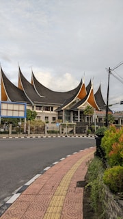 An architectural building with distinctively pointed, multi-tiered roofs resembling traditional Minangkabau architecture. It is situated at a corner with a curved road and a pedestrian pathway lined with vegetation. The sky is overcast with scattered clouds, and there are electrical wires visible.