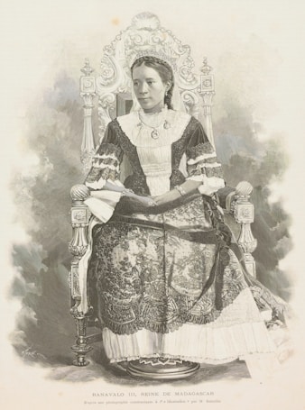 A woman in traditional attire sits on an intricately designed throne. She wears a dress adorned with lace and embroidery and a crown on her head, indicating royalty. Her expression is serene and dignified, exuding a sense of grace and authority.