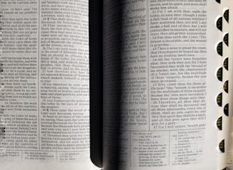An open book with visible text on both pages. The book seems to be religious, possibly a Bible, and includes chapter and verse numbers. The pages are divided into two columns with small font text. Rounded tabs along the right edge are marked with abbreviations of names, likely of books or chapters.