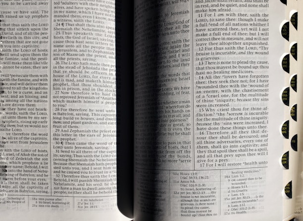 An open book with visible text on both pages. The book seems to be religious, possibly a Bible, and includes chapter and verse numbers. The pages are divided into two columns with small font text. Rounded tabs along the right edge are marked with abbreviations of names, likely of books or chapters.