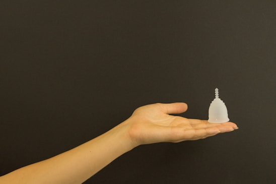 A human hand with an extended palm is holding a small, transparent menstrual cup against a plain, dark background.
